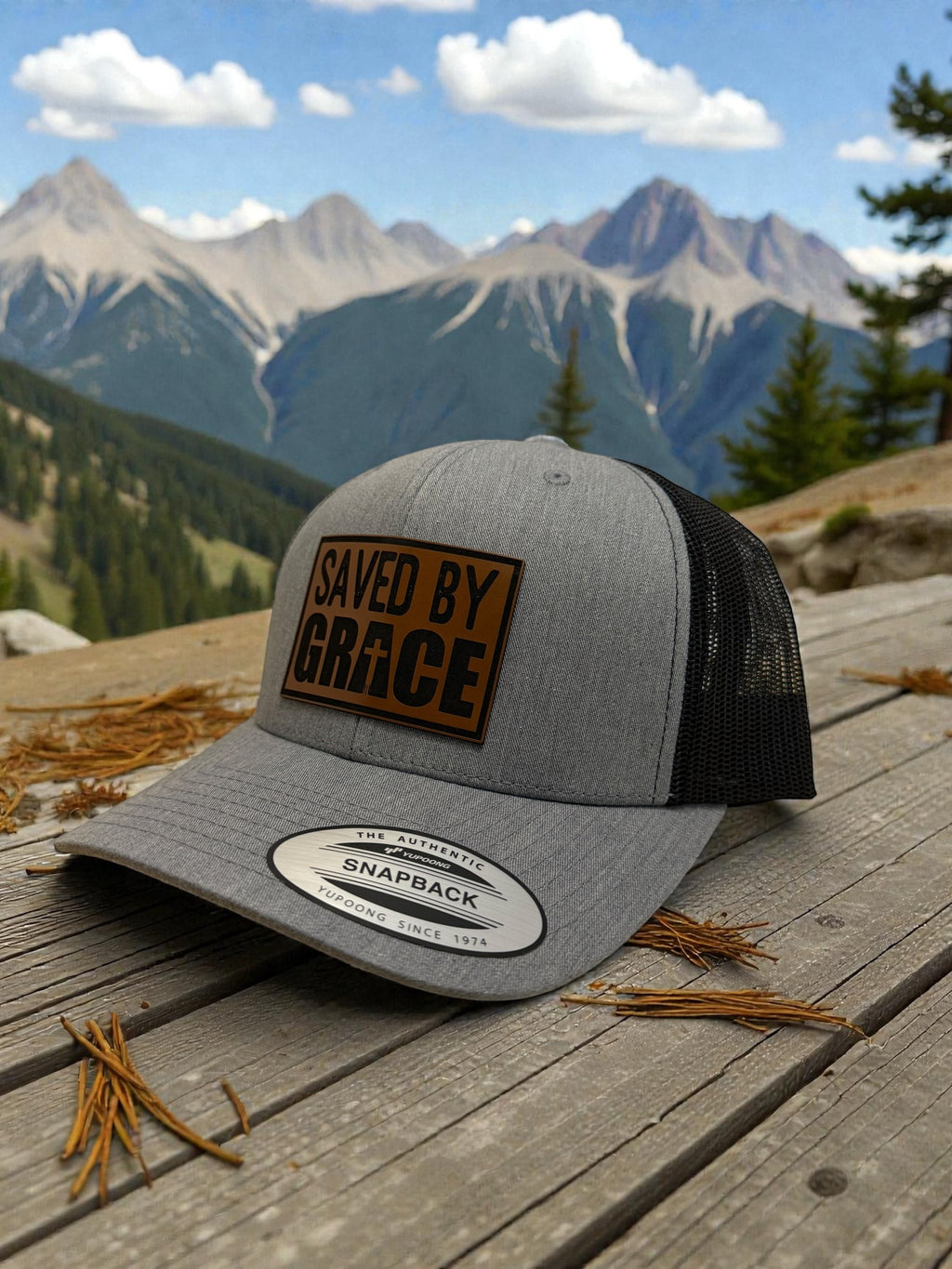 Gray cap with 'Saved by Grace' patch on a wooden surface with mountains in the background